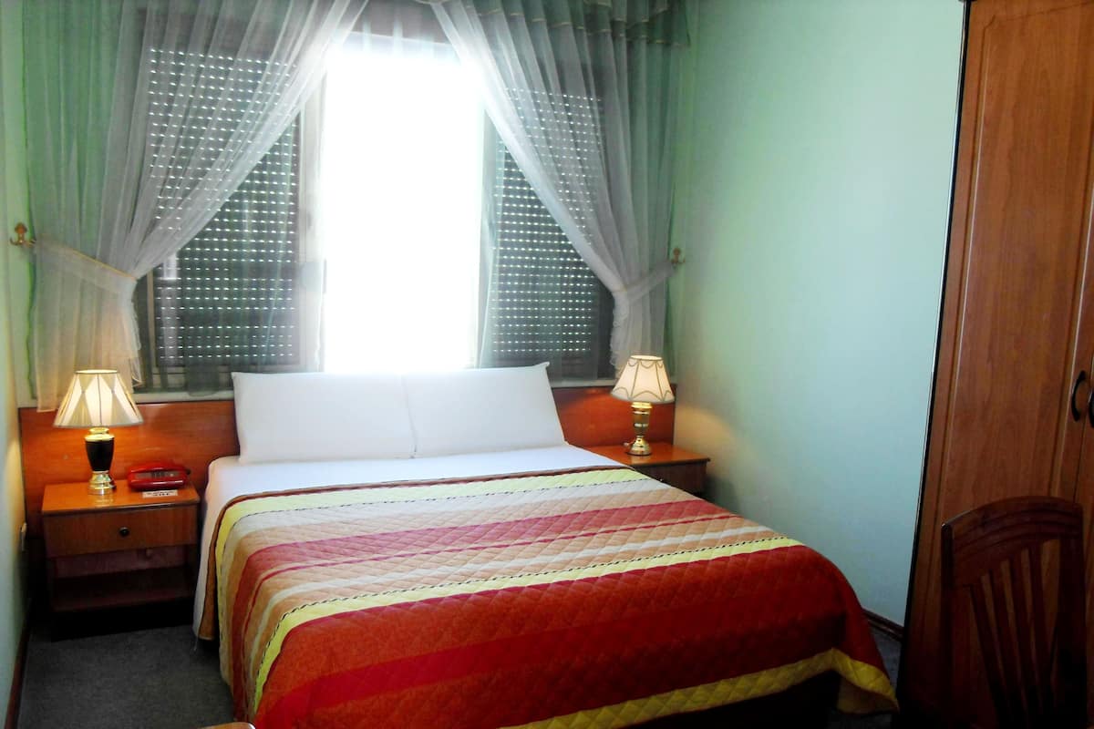 A hotel room with a bed, two bedside tables with lamps, a window with sheer curtains, and a wooden wardrobe.
