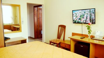 A hotel room with a bed, a wooden chair, a desk with a vase of flowers, a television, and a mirror.