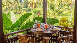 Una mesa de madera dispuesta con cosas para el desayuno, incluyendo una tetera, tazas y un plato de comida, en un balcón con vista a vegetación exuberante y palmeras.