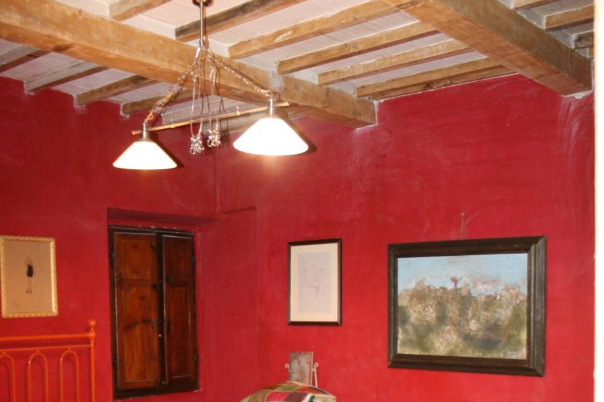 Une pièce avec un mur rouge, un lit recouvert d’une courtepointe colorée, une commode en bois et un lustre.