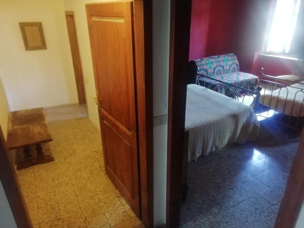 A room with a wooden door, a bench, a bed with a patterned bedspread, and a chair with a floral cushion.