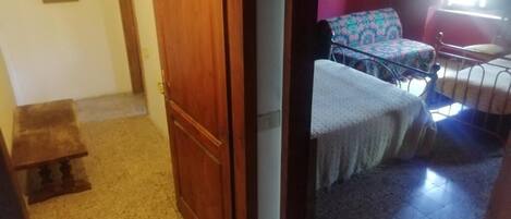 Una habitación con puerta de madera, un banco, una cama con cubrecama estampado y una silla con cojín floral.