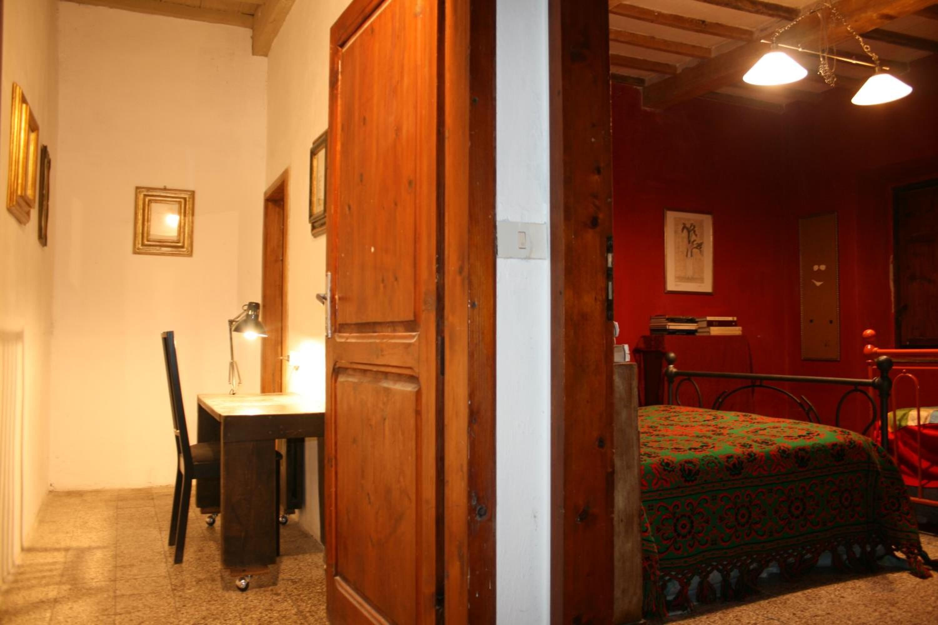 A room with a wooden door, a desk with a chair, a bed with a patterned bedspread, and a red wall with framed pictures.