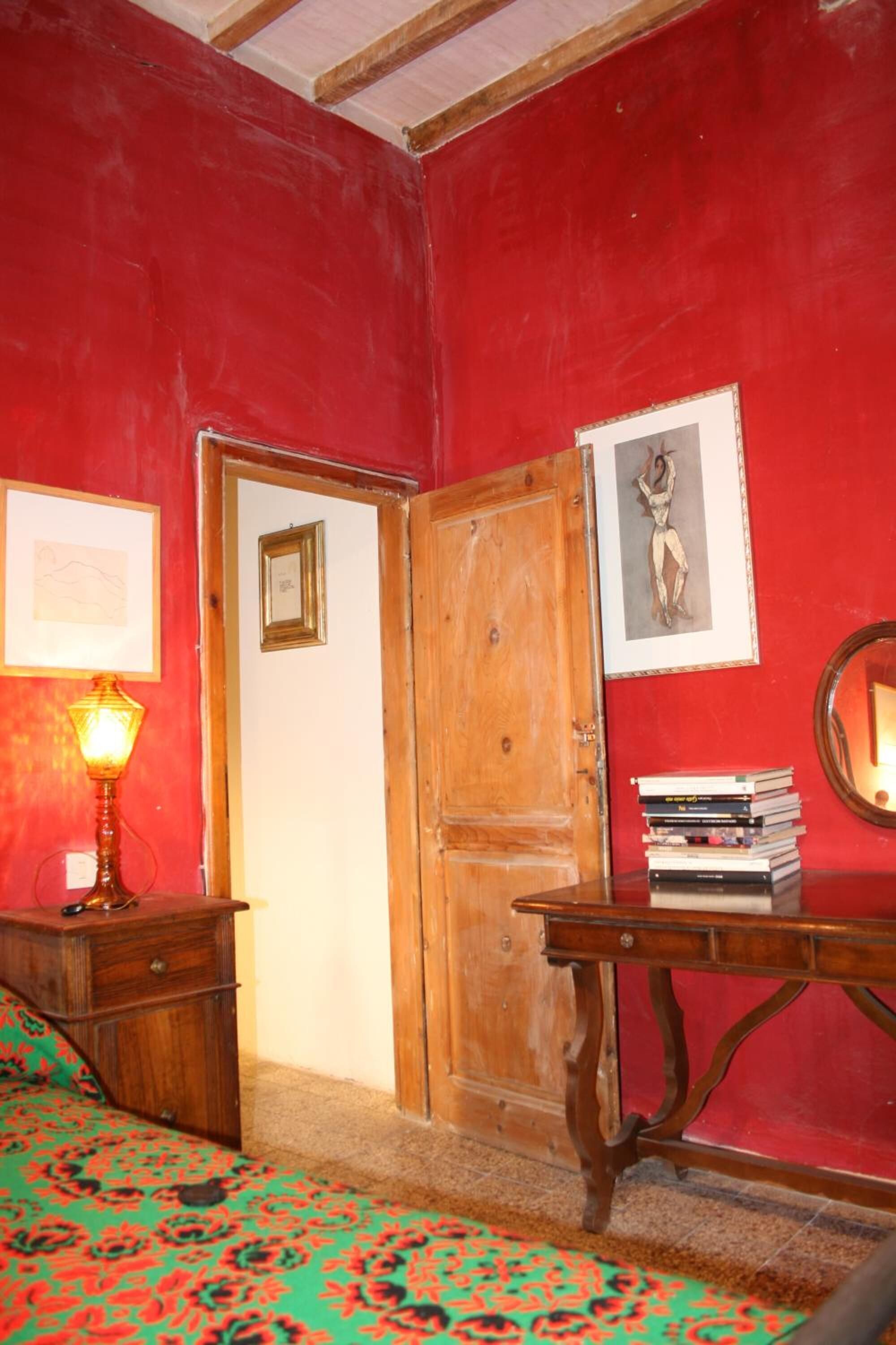 Una habitación con paredes rojas, una puerta de madera, una cama con colcha de cuadros verdes y rojos, una mesita de noche de madera con una lámpara, una mesa con libros y un cuadro enmarcado en la pared.