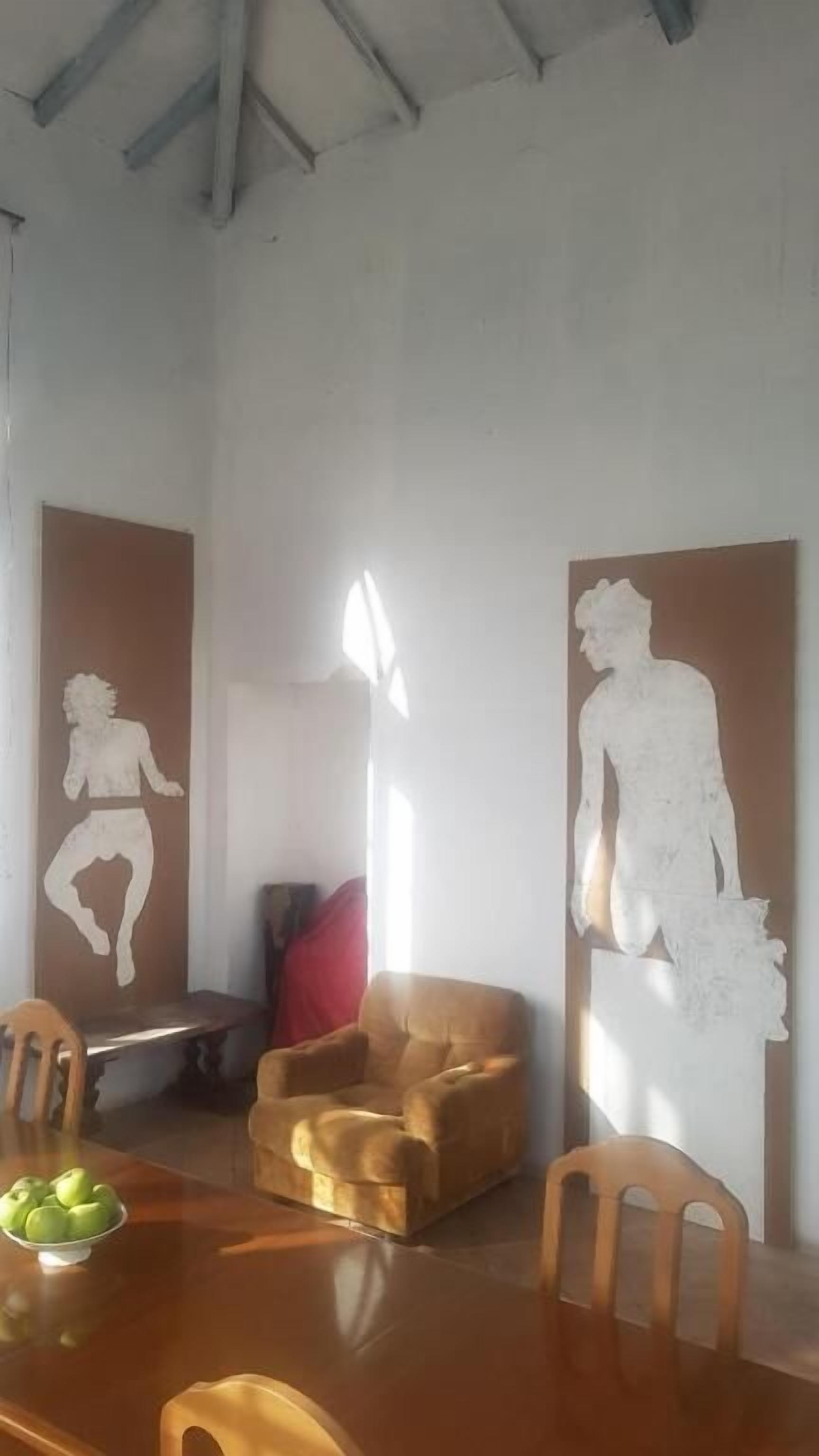 Zona pranzo con un tavolo in legno, sedie e una ciotola di frutta. Sono presenti opere d'arte a parete e un cuscino rosso sul tavolo.