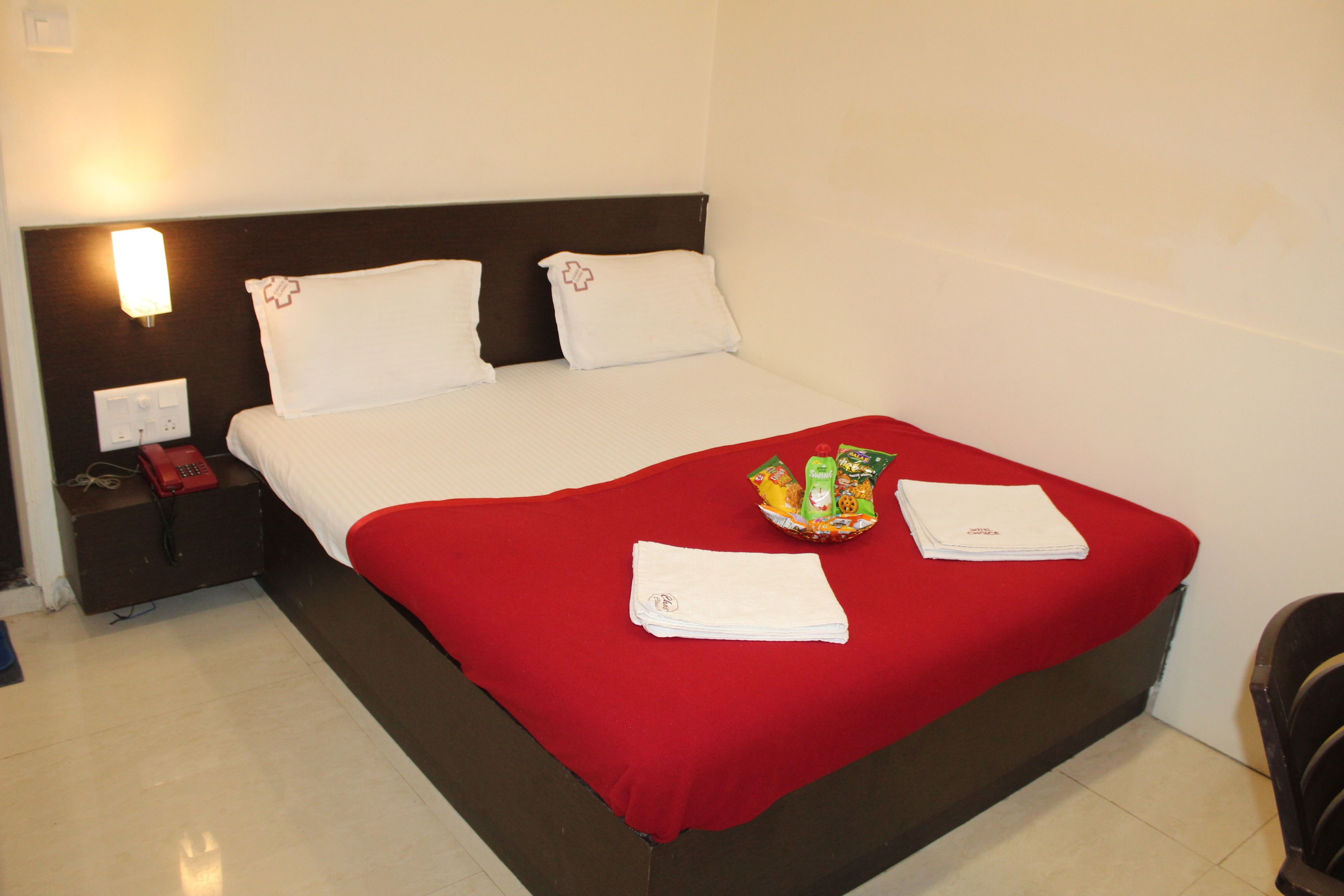 A hotel room with a bed, a red blanket, a nightstand with a phone, and a basket of toiletries.