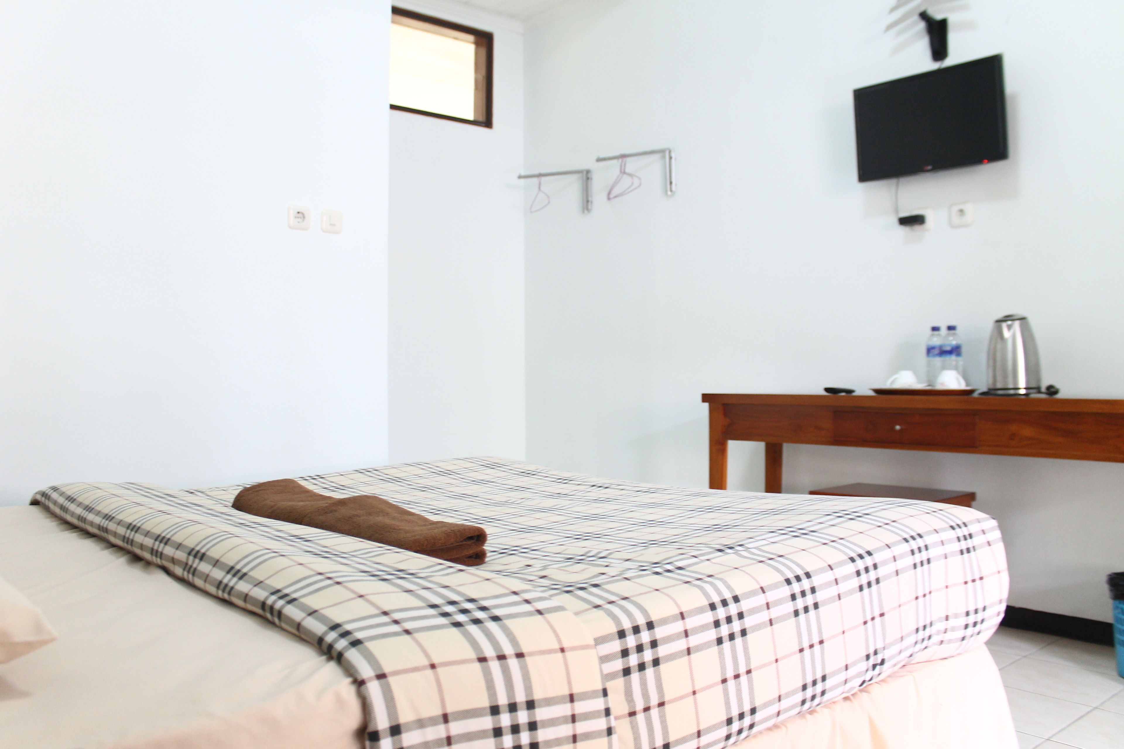 A bedroom with a bed, a TV mounted on the wall, a desk with a kettle, and a trash bin.