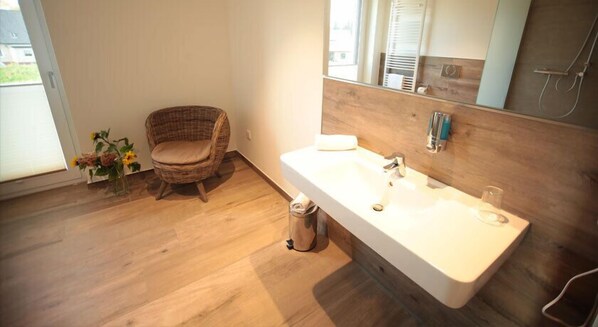 Un baño con piso de madera, una silla de mimbre, un lavamanos blanco y un espejo.