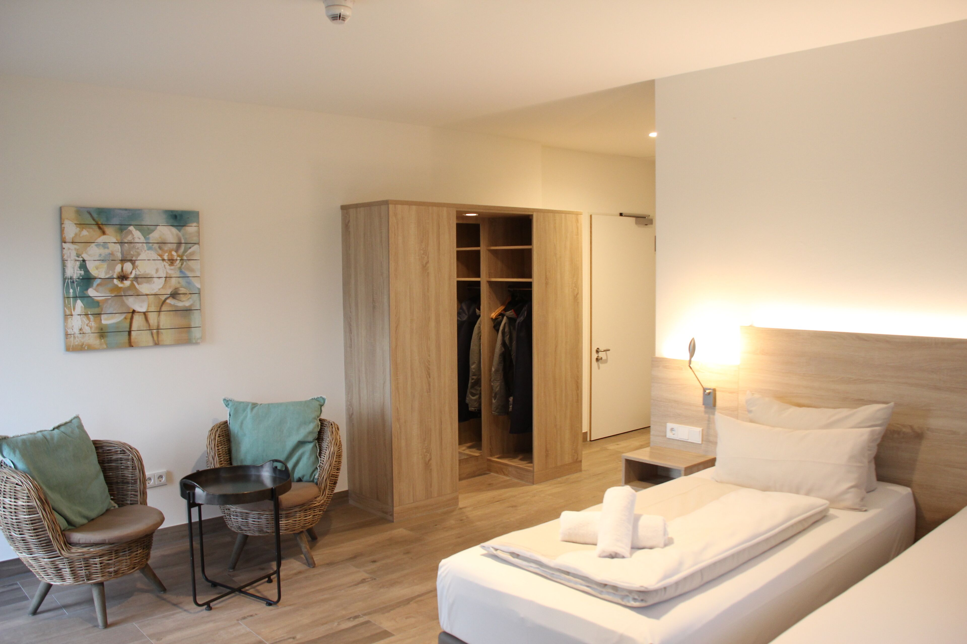 A bedroom with a bed, a wooden wardrobe, a wicker chair, a small table, and a wall-mounted artwork.