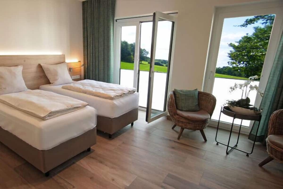 Habitación doble con dos camas, una silla y vista a un paisaje verde a través de un ventanal.