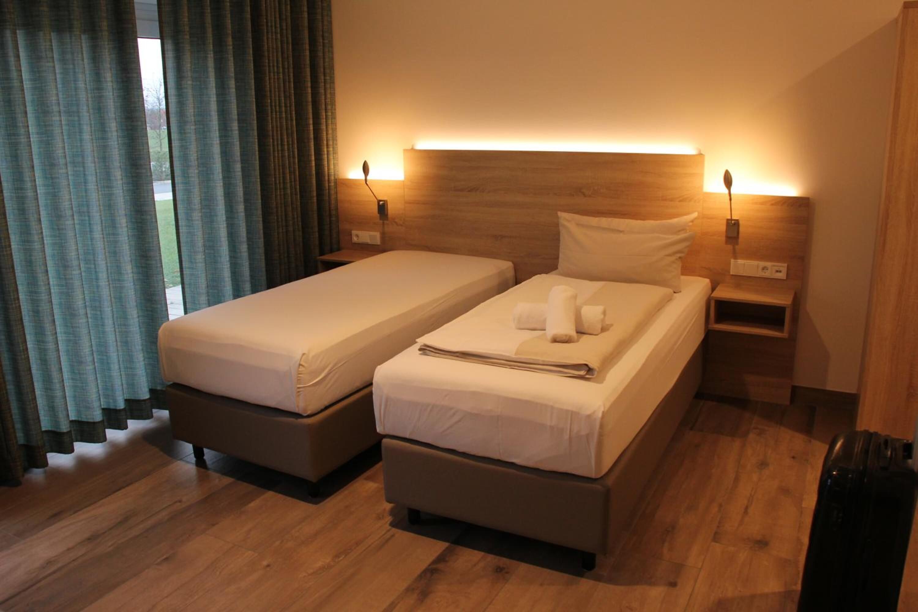A double room with two beds, wooden headboards, bedside lamps, and a view of the outdoors through curtains.