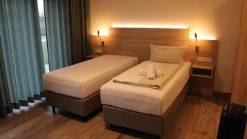 A double room with two beds, wooden headboards, bedside lamps, and a view of the outdoors through curtains.