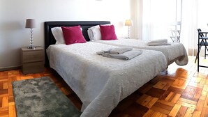 A double bed with white bedding and two pillows, a nightstand with a lamp, a green rug, and a wooden floor. - Apartamento À Cedofeita (Porto)