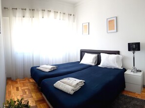 A hotel room with two beds, a nightstand, a lamp, and a window with sheer curtains. - Apartamento À Cedofeita (Porto)