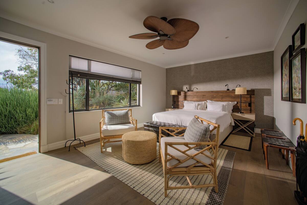 A modern bedroom with a large bed, two chairs, a small round table, a ceiling fan, and a view of the outdoors.