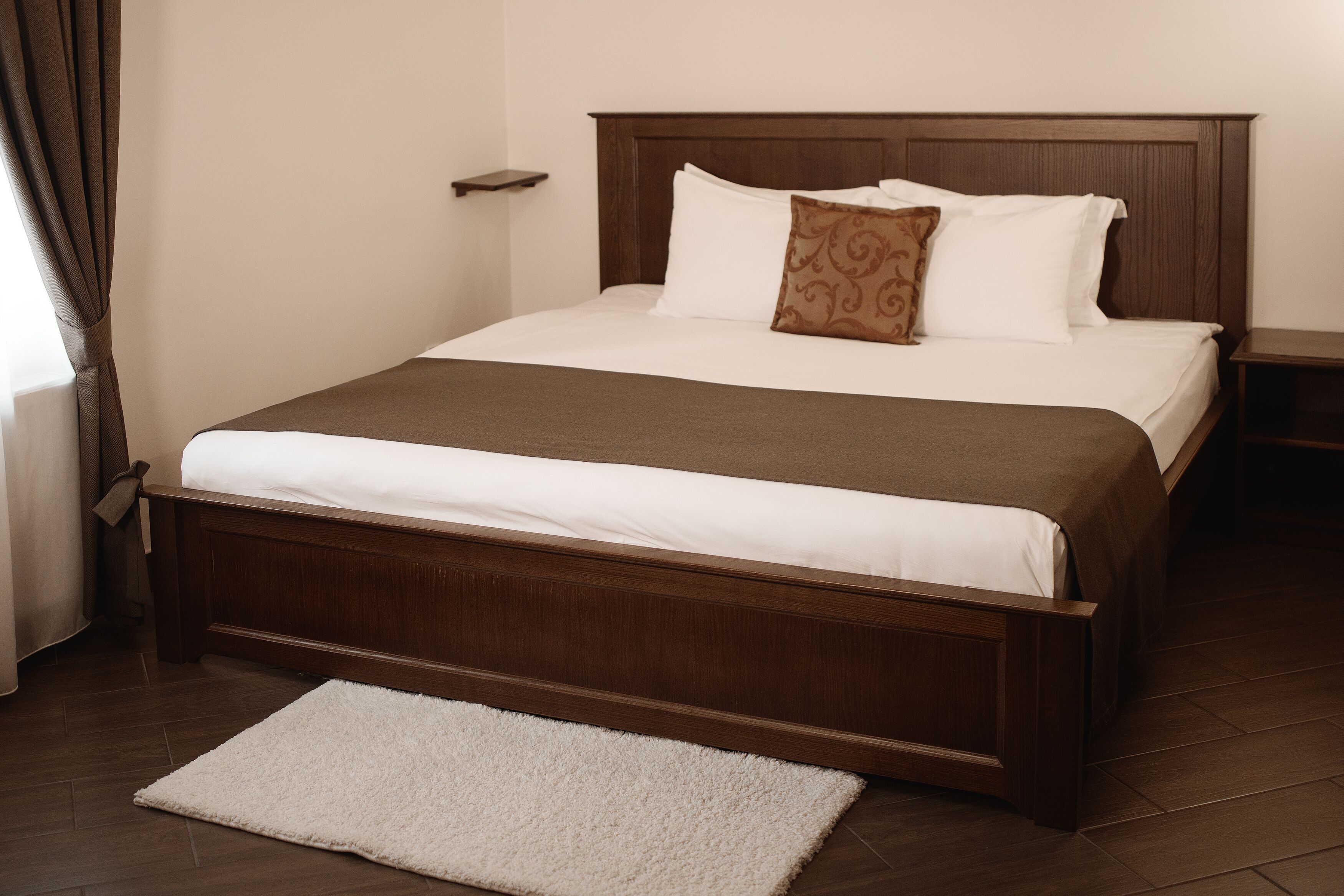 Una cama bien hecha con cabecera de madera, manta blanca y marrón, y alfombra blanca en el suelo.