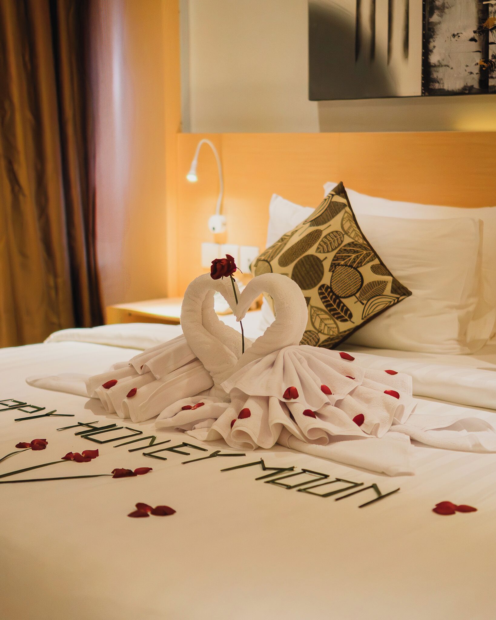 A neatly made bed with a heart-shaped towel arrangement, a rose, and a pillow with a leaf pattern.