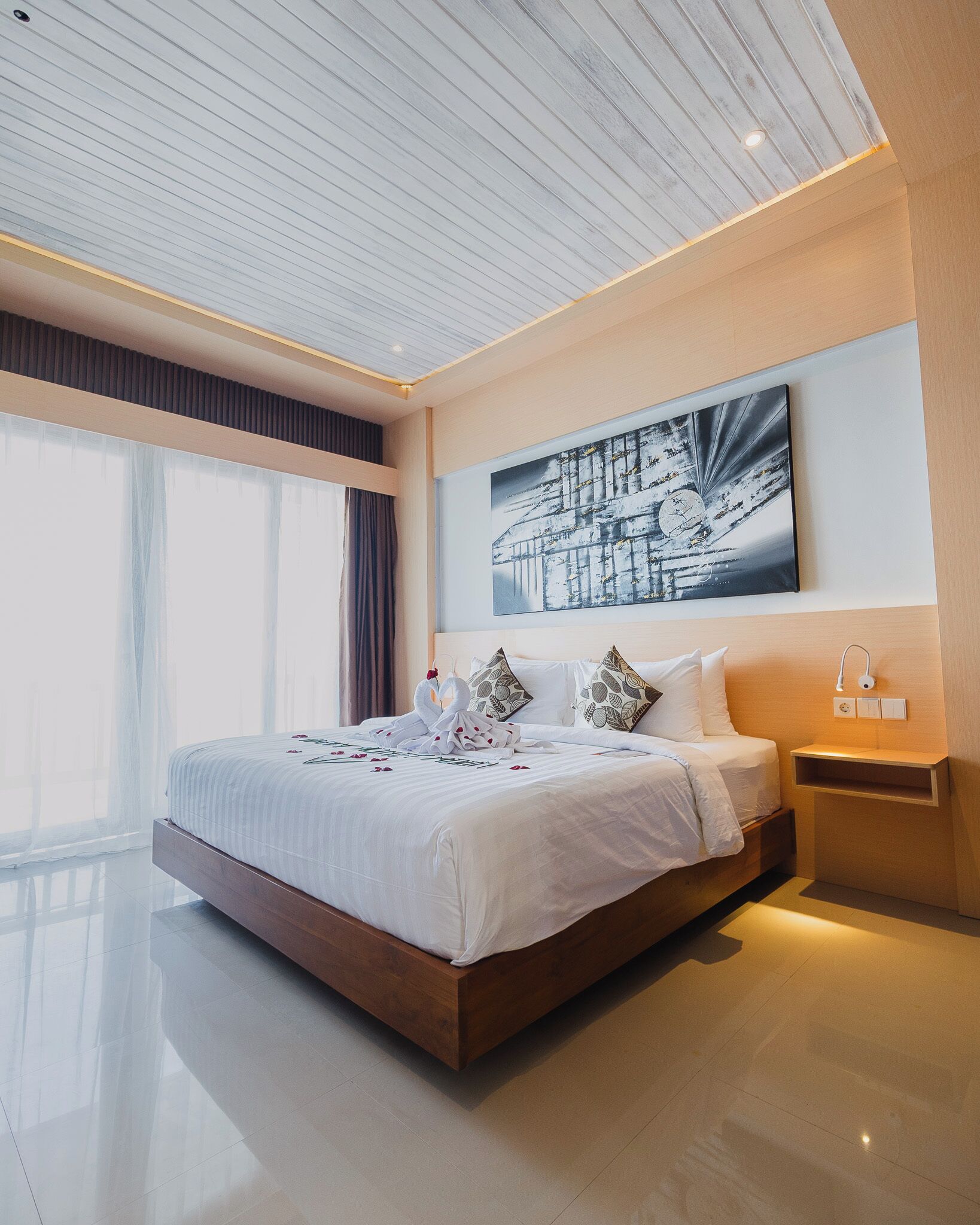 A modern bedroom with a large bed, a nightstand, a wall-mounted light, and a large window with curtains.