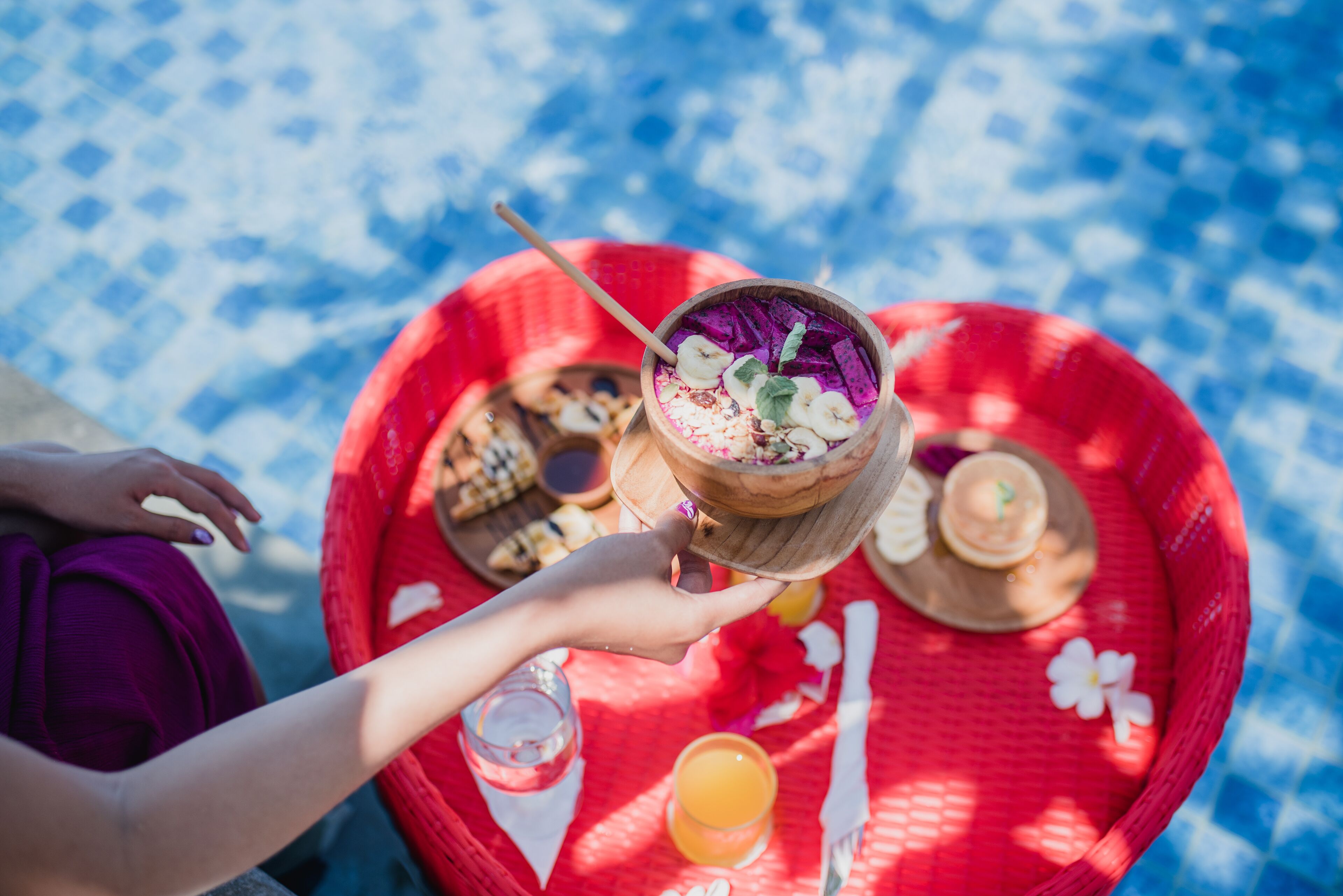 A person holding a wooden bowl with a purple drink, a red tray with food and drinks, and a blue patterned tablecloth.