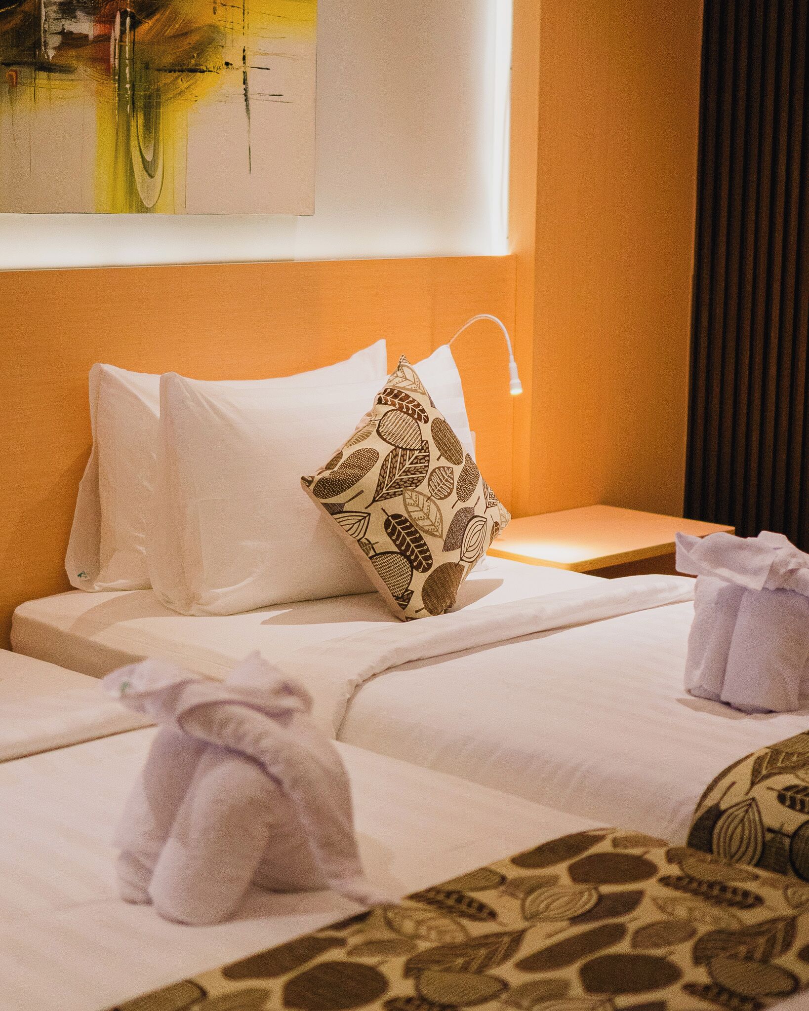 A hotel room with two beds, white linens, a patterned pillow, a bedside table with a lamp, and a painting on the wall.