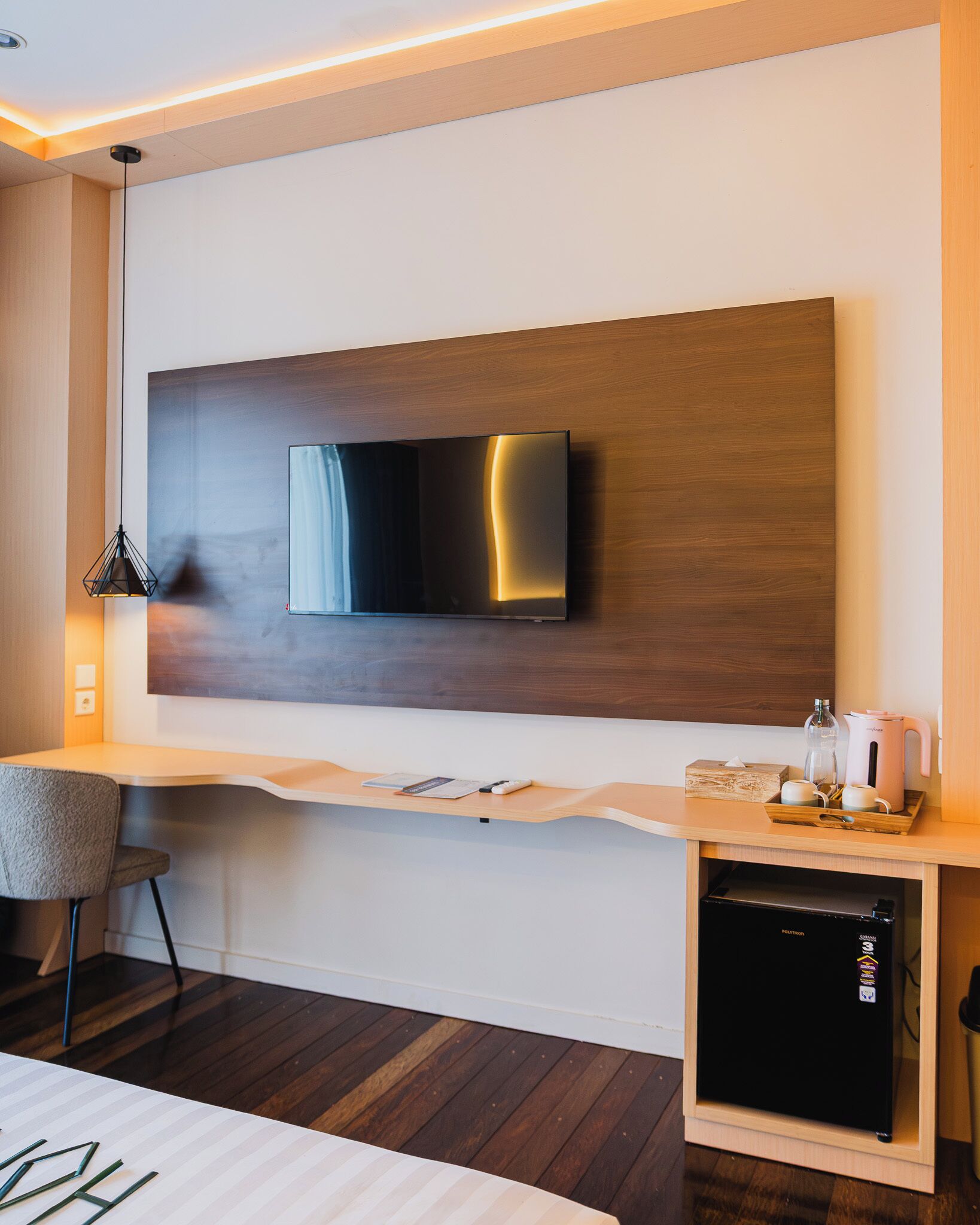 Une chambre d’hôtel moderne équipée d’un téléviseur à écran plat fixé sur un panneau en bois, d’un petit bureau avec une chaise, d’une machine à café et d’un mini-réfrigérateur.