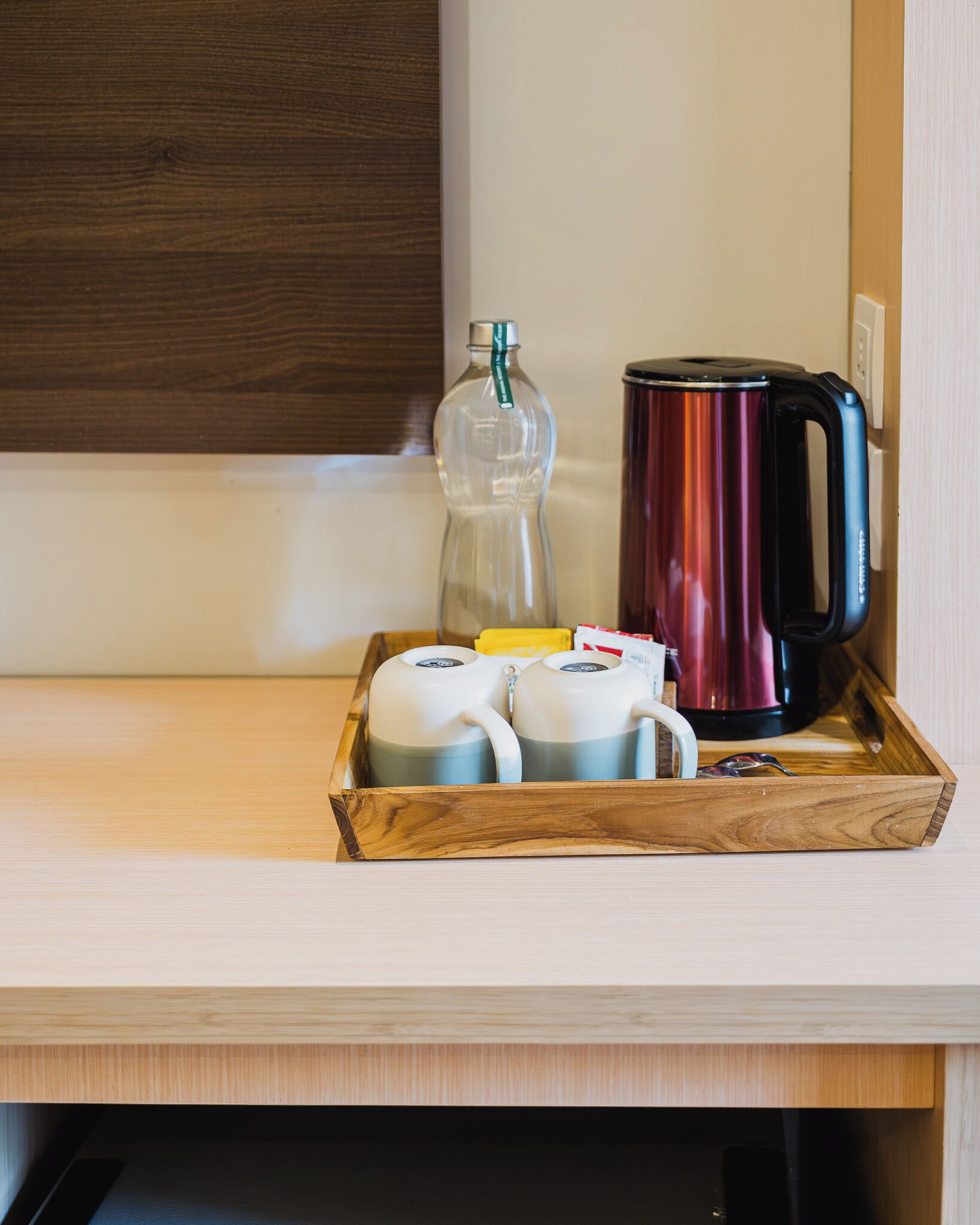 Een houten dienblad met een glazen fles, een zwart en rood elektrisch waterkoker en twee witte mokken op een houten tafel.