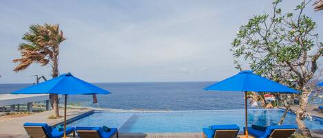 Um deck de madeira com espreguiçadeiras e guarda-sóis azuis, com vista para uma piscina e o oceano.
