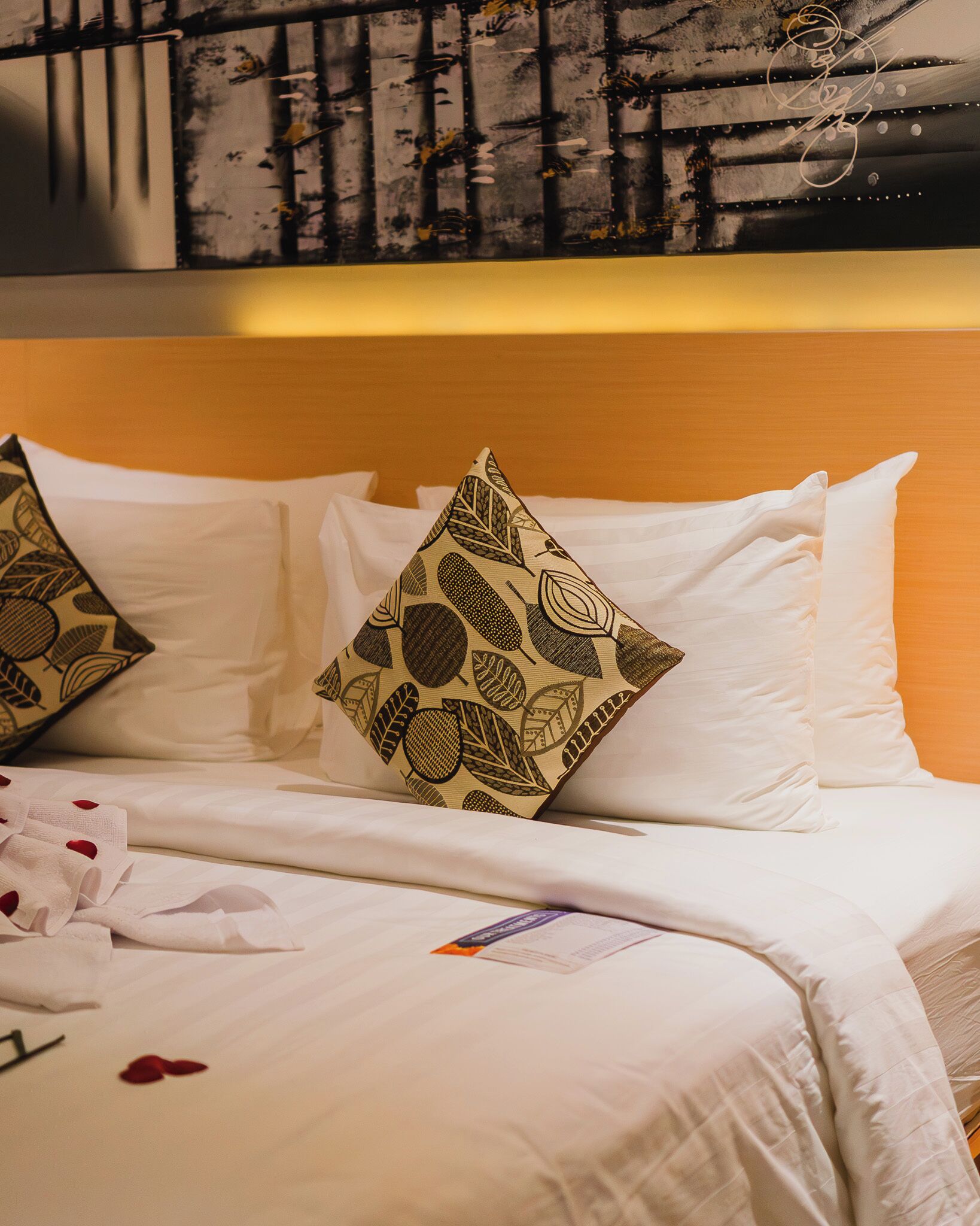 A neatly made bed with white linens and patterned pillows, a wooden headboard, and a wall with a textured mural.