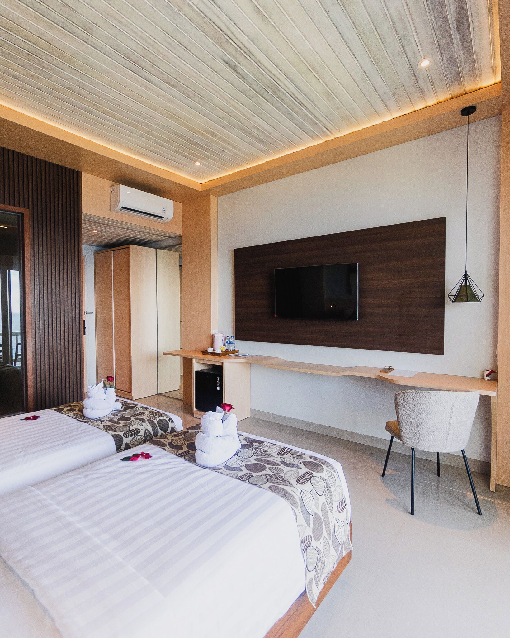 Ein modernes Hotelzimmer mit zwei Betten, einem an einer Holzplatte montierten Fernseher, einem Schreibtisch mit Stuhl und einer Klimaanlage.