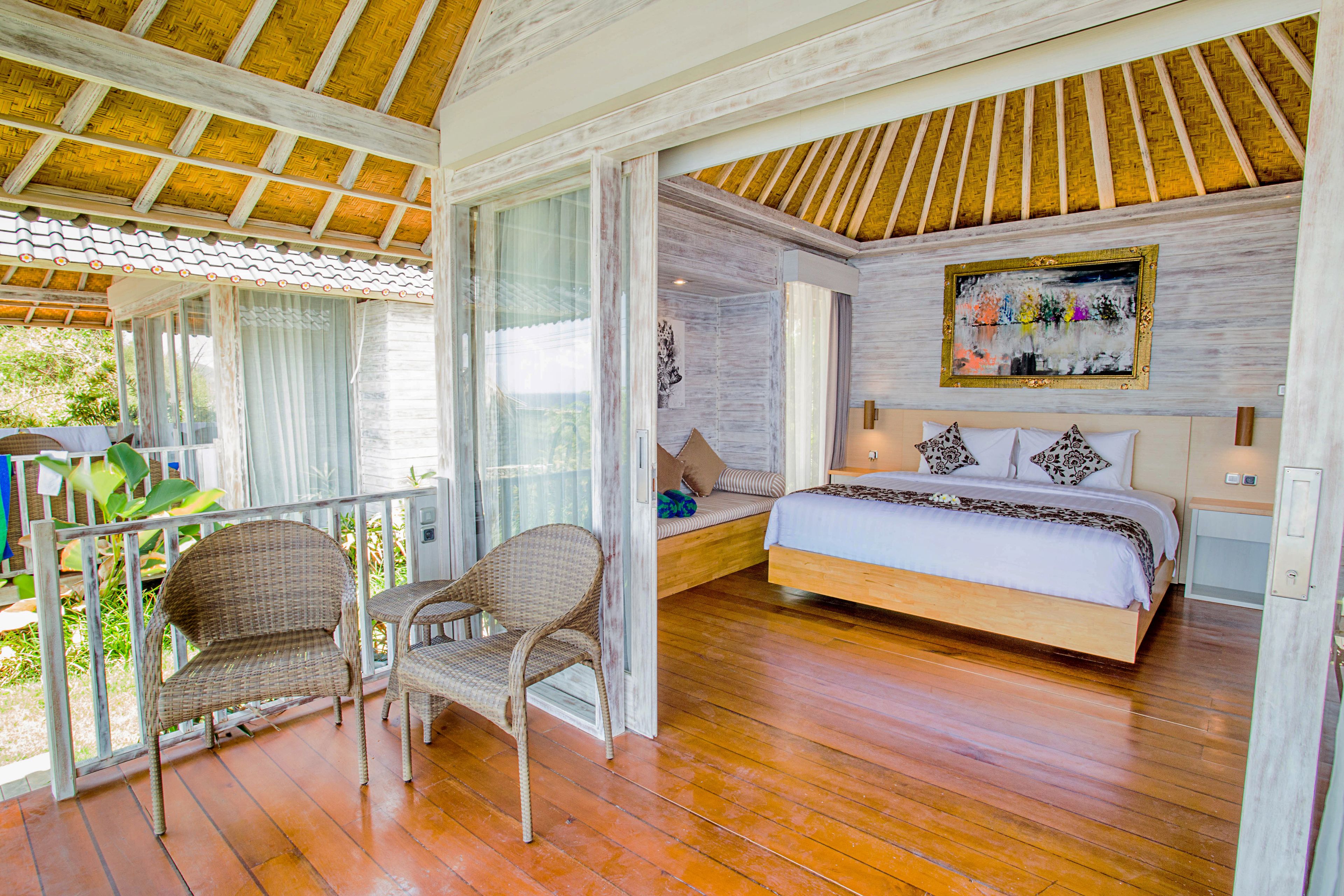 A bedroom with a wooden floor, a bed with a white and patterned bedspread, two wicker chairs, and a large window with curtains.