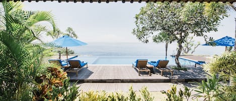 Eine Holzterrasse mit Liegestühlen und Sonnenschirmen mit Blick auf ein Schwimmbecken und das Meer.