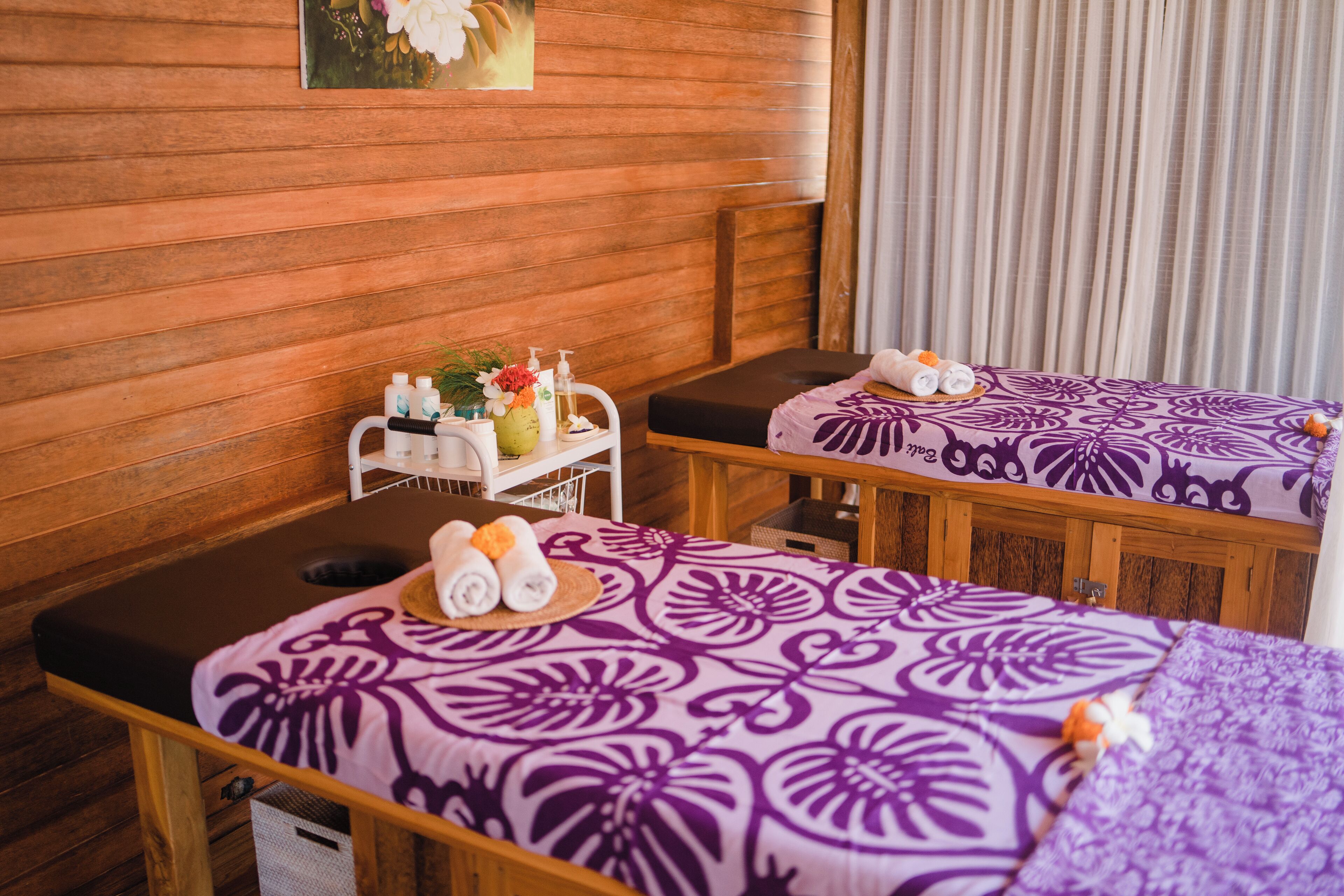 A spa room with wooden walls, two massage tables with purple floral bedspreads, a small table with toiletries, and a painting on the wall.