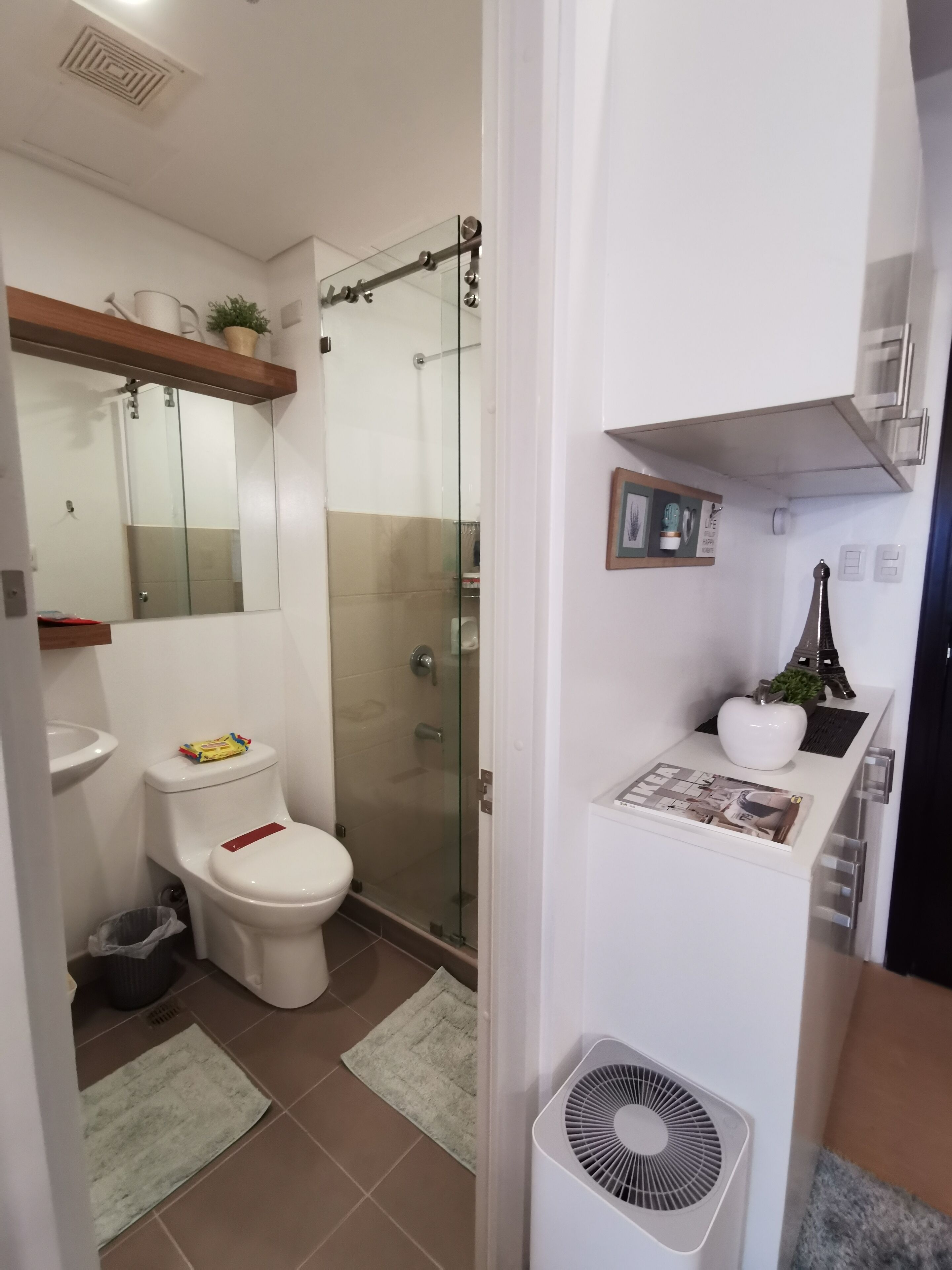 Une salle de bain comprenant des toilettes, une douche et un espace de douche fermé par des parois en verre. On y trouve un lavabo blanc, un miroir et une petite étagère avec des objets décoratifs.