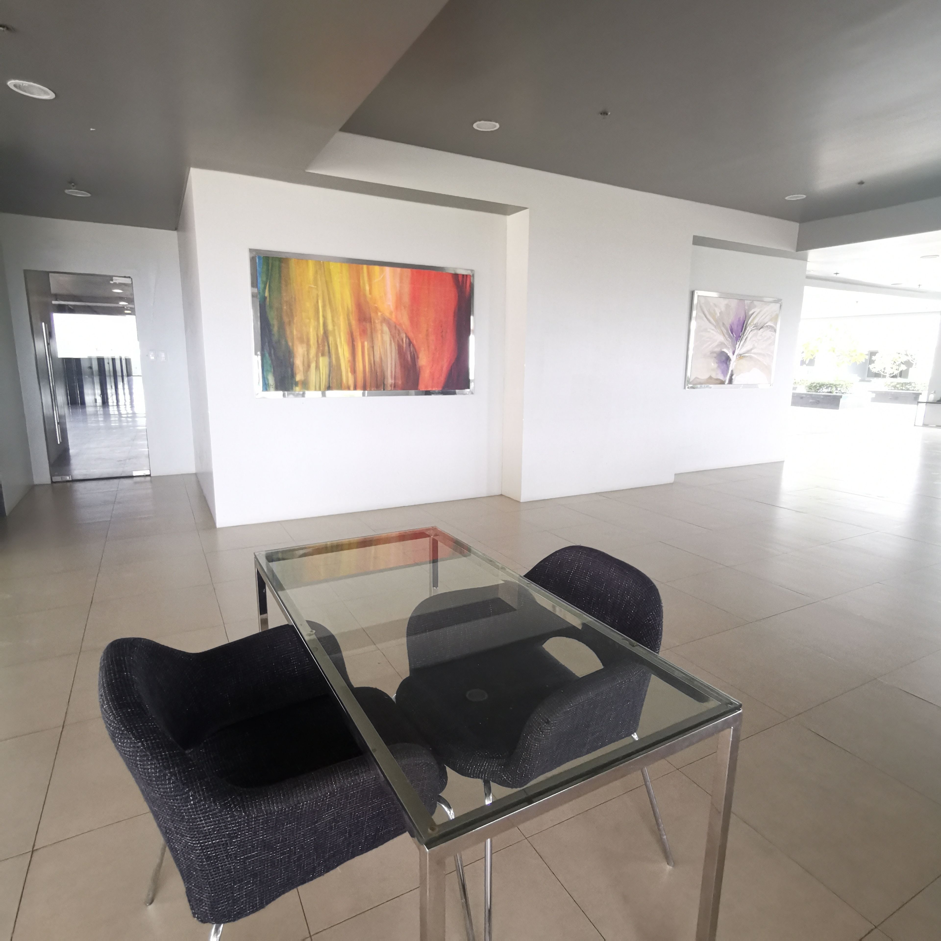 A modern meeting room with a glass table and two chairs, a colorful abstract painting on the wall, and a view of a garden through large windows.