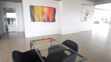 A modern meeting room with a glass table and two chairs, a colorful abstract painting on the wall, and a view of a garden through large windows.