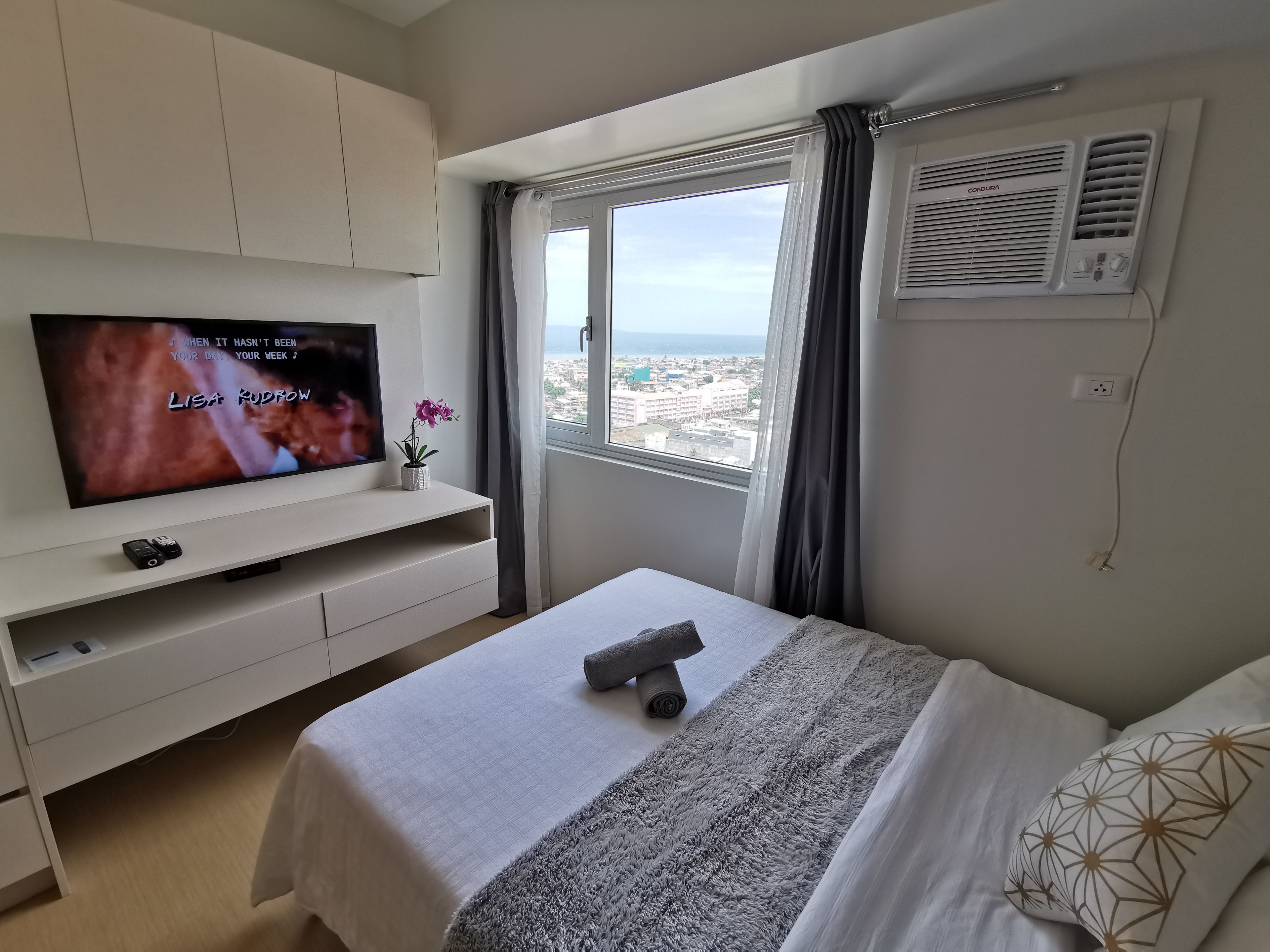 Een hotelkamer met een bed, een televisie op een wit kastje, een raam met uitzicht op de stad, en een airconditioner.