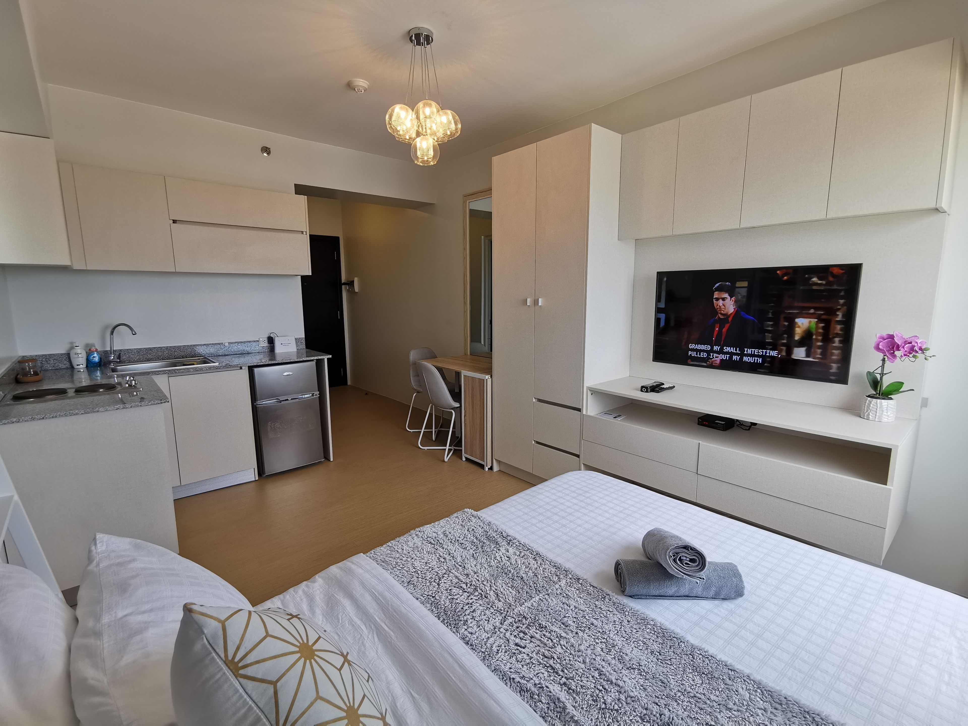 A modern hotel room with a kitchenette, a TV, a bed with a rolled-up towel, and a small desk area.