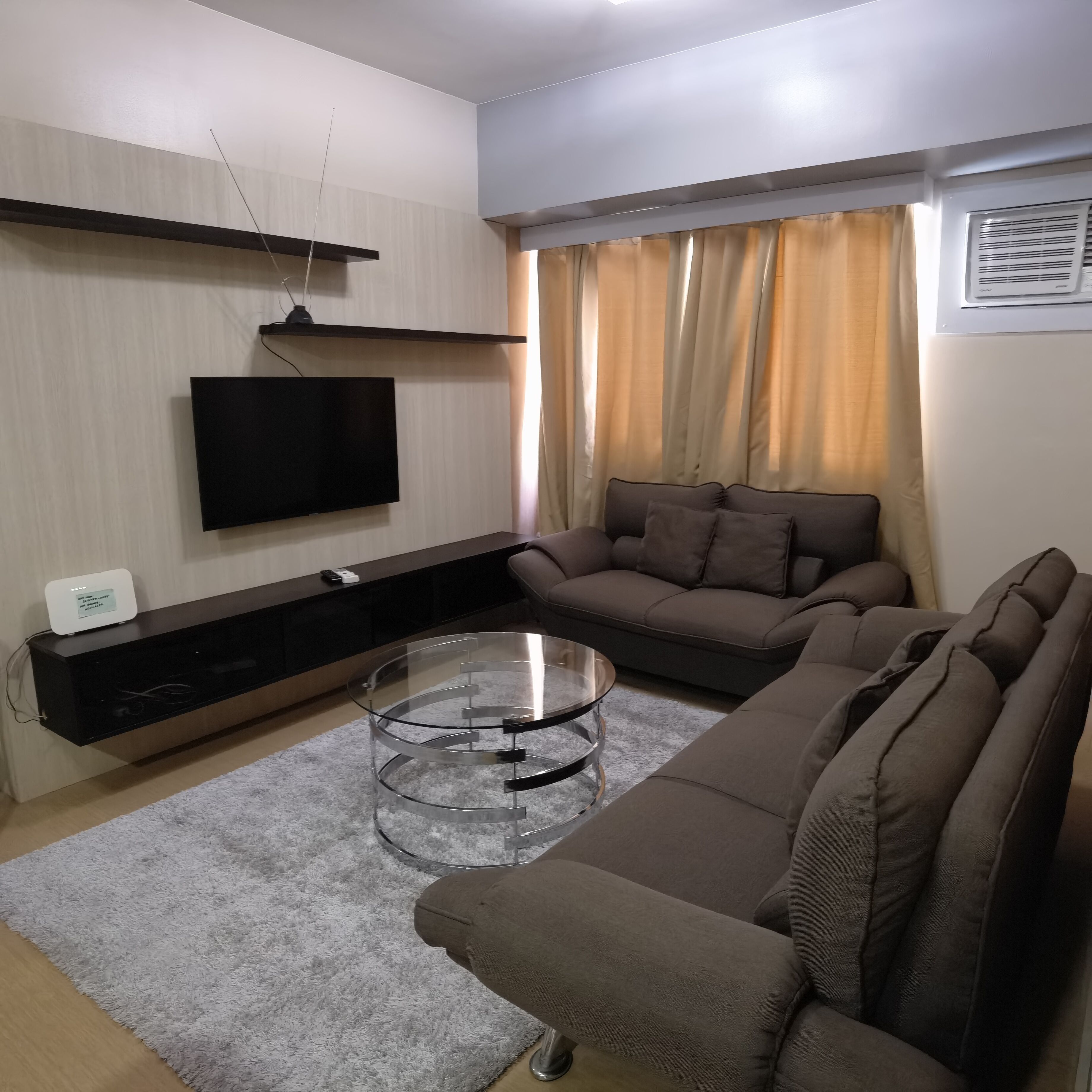A modern living room with a flat-screen TV mounted on the wall, a dark wooden entertainment unit, a brown sofa, and a glass coffee table.