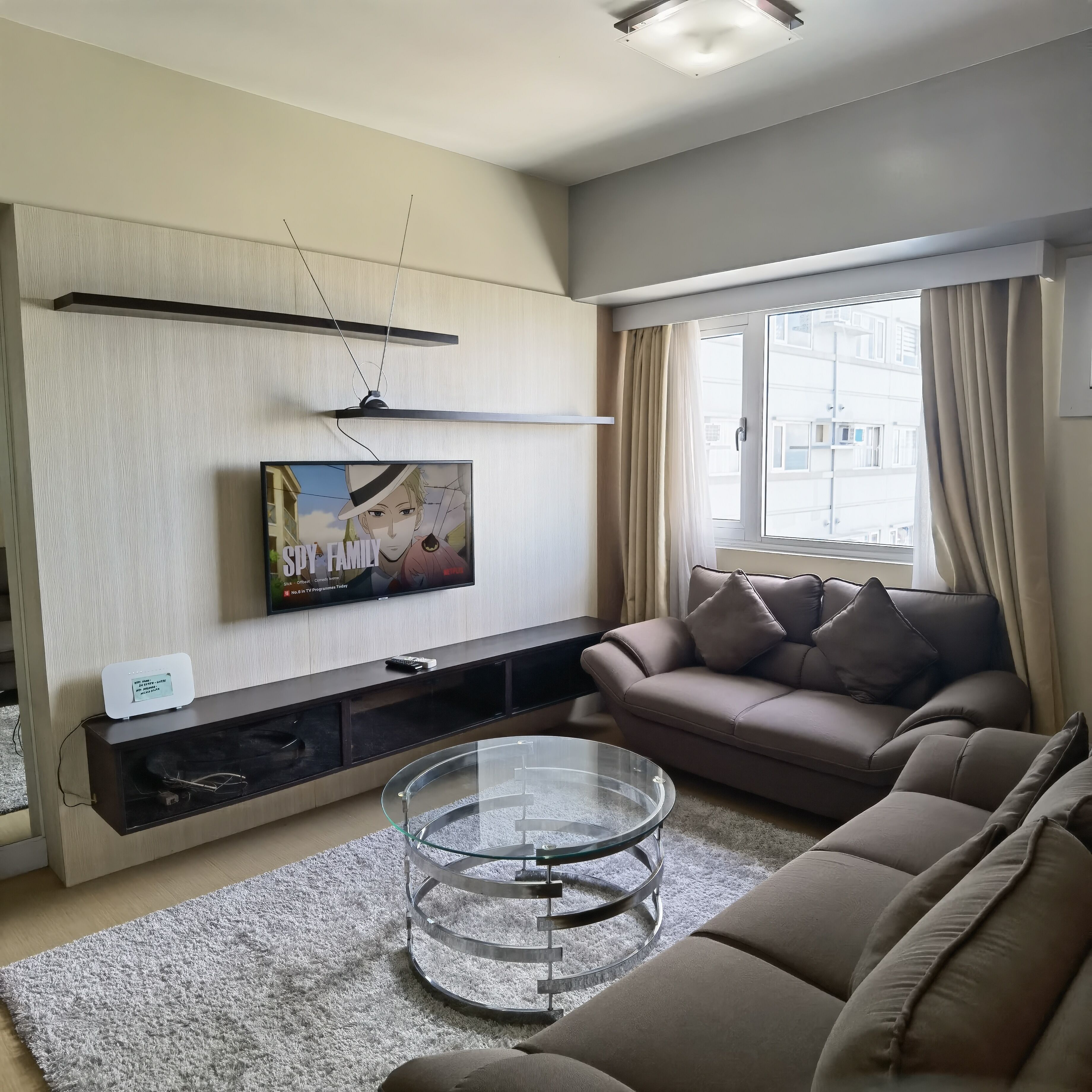 A modern living room with a flat-screen TV, a sofa, a glass coffee table, and a large window with curtains.