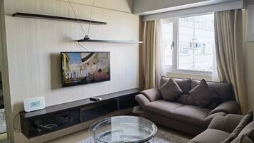 A modern living room with a flat-screen TV, a sofa, a glass coffee table, and a large window with curtains.