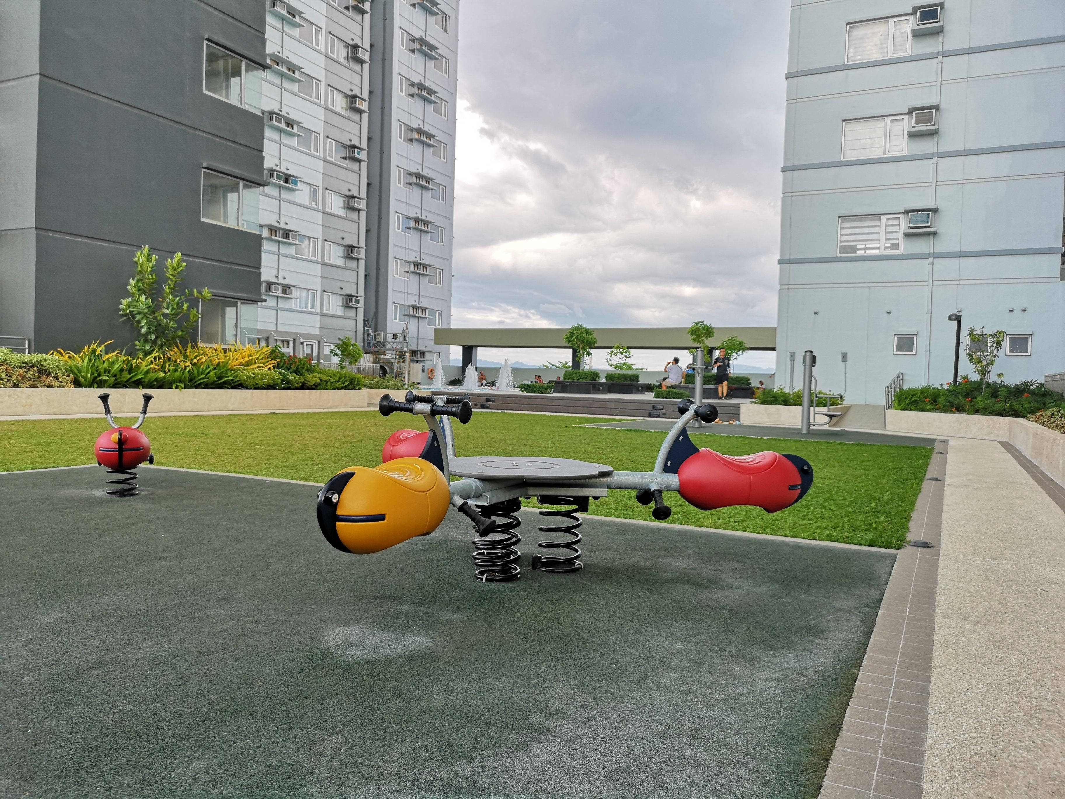 Un gimnasio al aire libre con equipamiento de ejercicio, rodeado de un área ajardinada y edificios modernos al fondo.