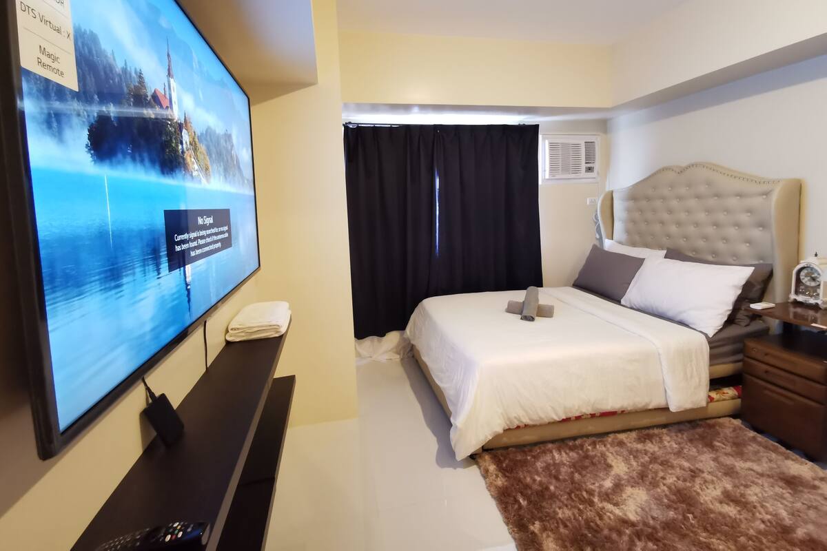 Hotellihuone, jossa on suuri, seinälle asennettu taulutelevisio, valkoisilla lakanoiden peitetty sänky, ruskea matto, yöpöytä lampun kanssa ja ikkuna, jossa on mustat verhot.