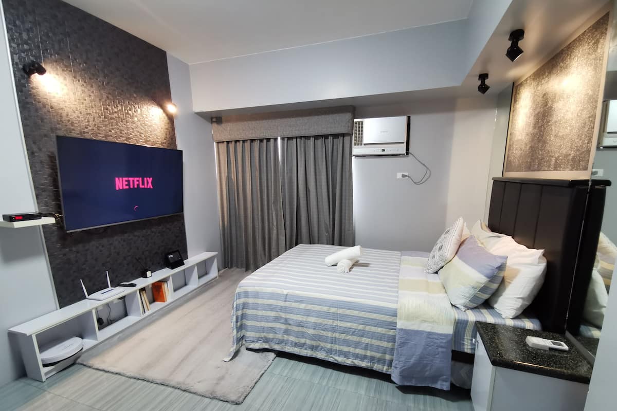 Moderni hotellihuone, jossa on suuri, litteänäyttöinen televisio, josta voi katsoa Netflixiä, raidallinen vuodevaatteilla varustettu sänky, yöpöytä, pieni työpöytä lampun kanssa ja ikkuna, jossa on verhot.