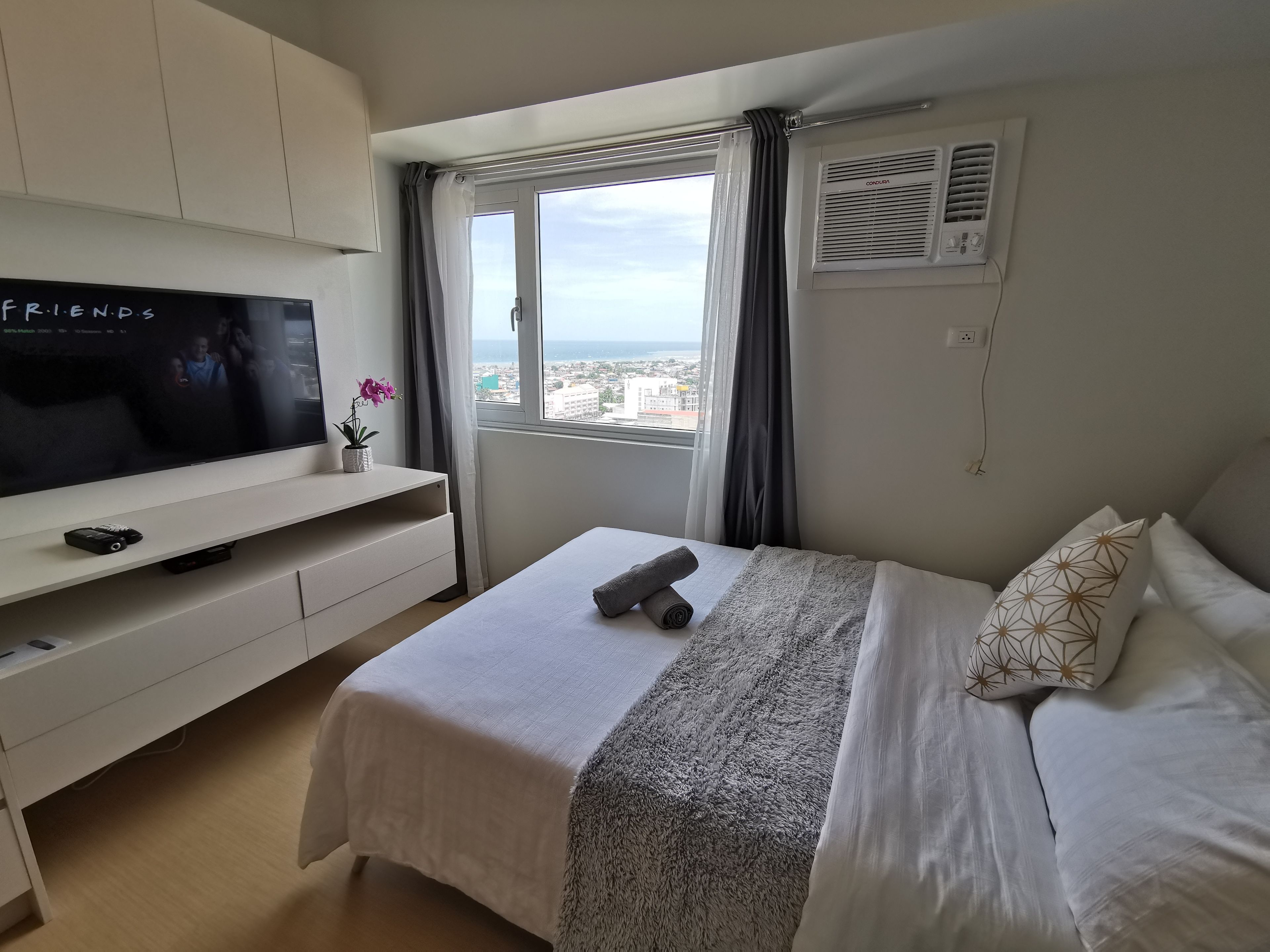 A hotel room with a bed, a TV on a white cabinet, a window with curtains, and an air conditioning unit.
