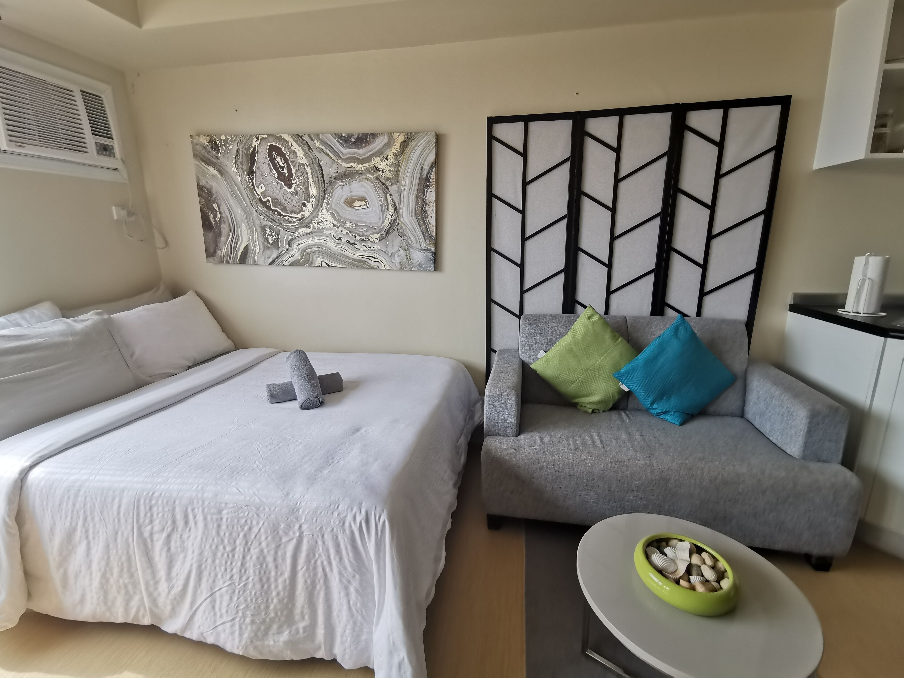 A hotel room with a bed, a gray sofa, a decorative wall panel, and a small table with a bowl.