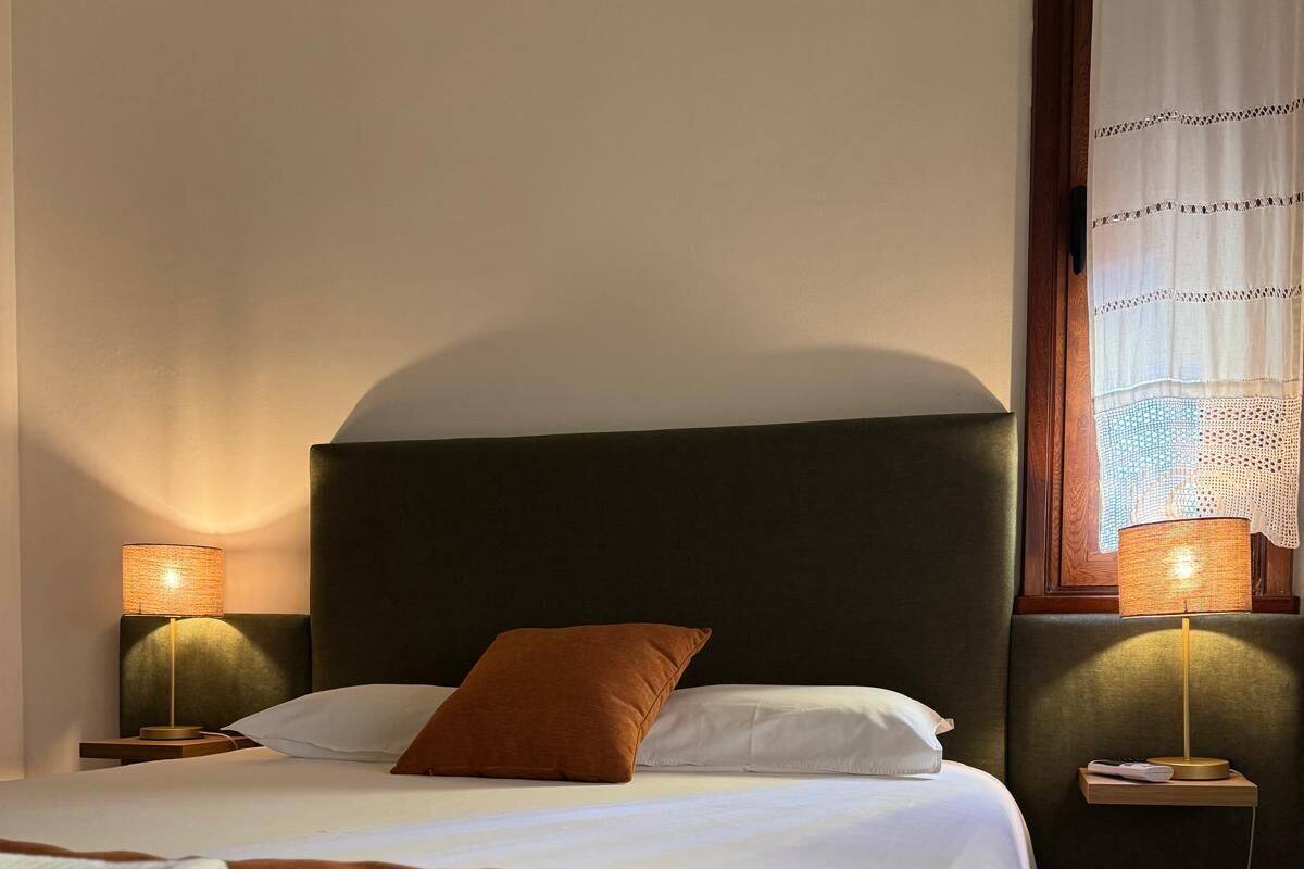 Una cama bien hecha con una manta marrón, dos lámparas de noche y una ventana con cortina.