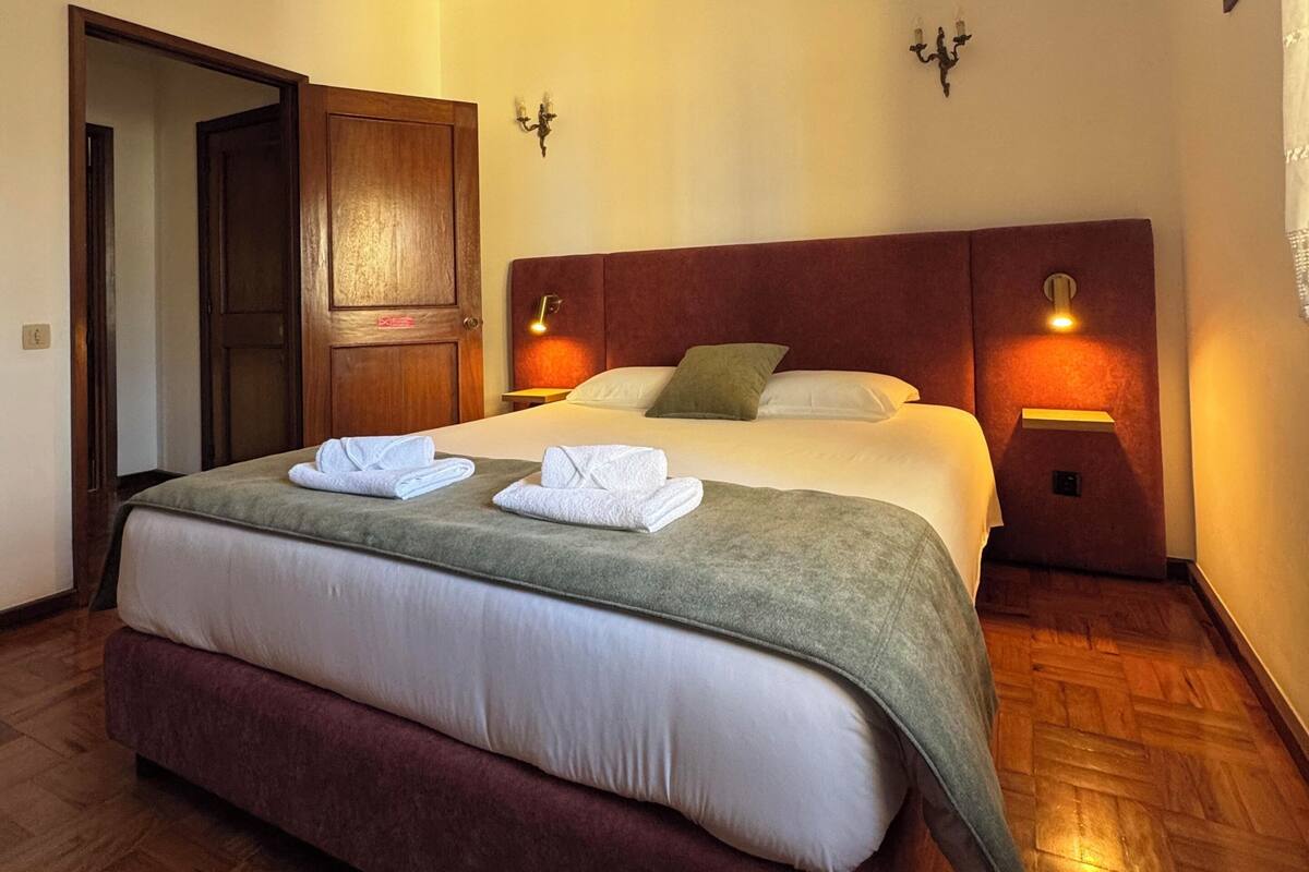 Una habitación de hotel con una cama grande, dos mesitas de noche, un armario de madera y una puerta que da a otra habitación.