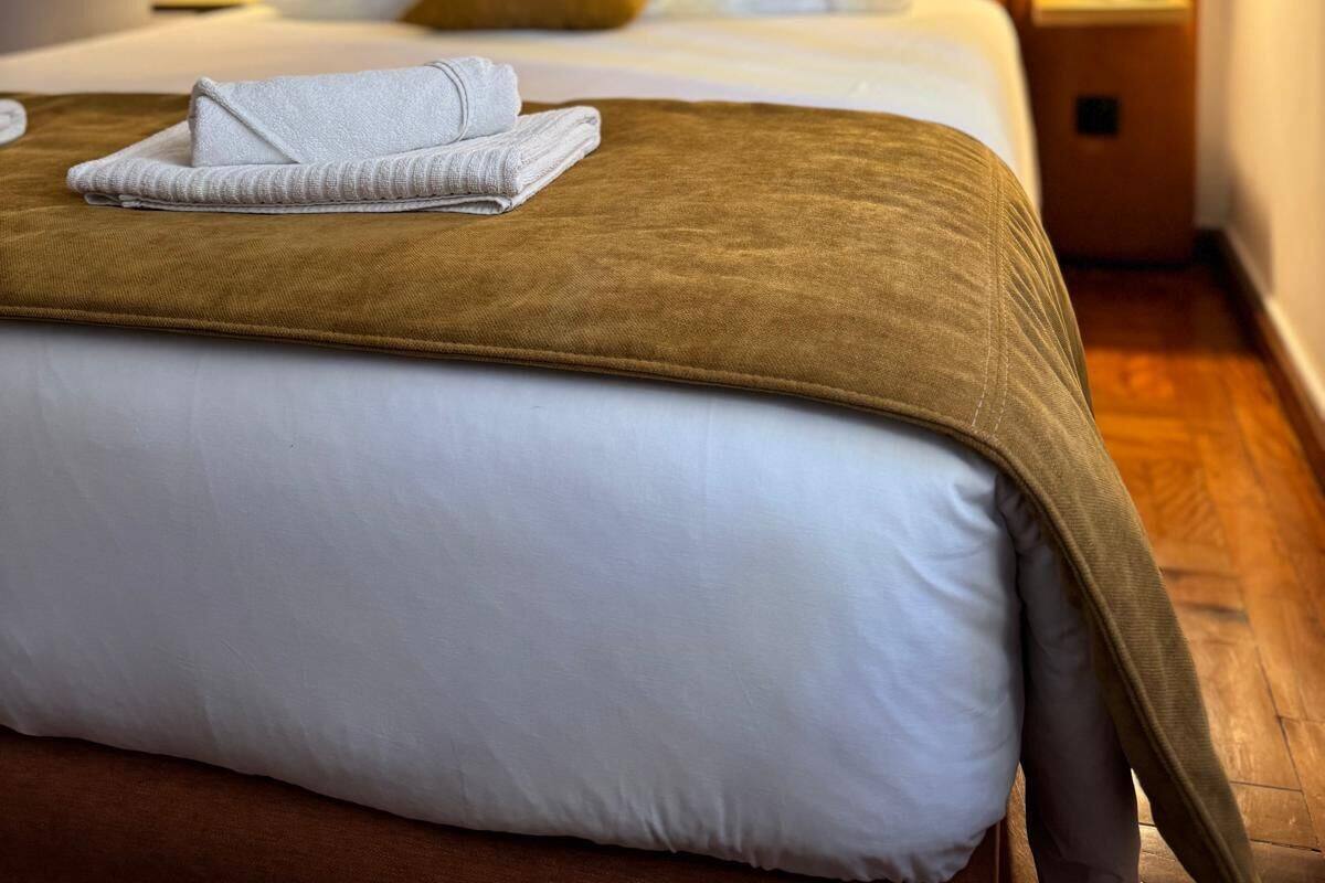 Una cama bien hecha con cabecera marrón, ropa de cama blanca y una toalla doblada en el pie de cama.