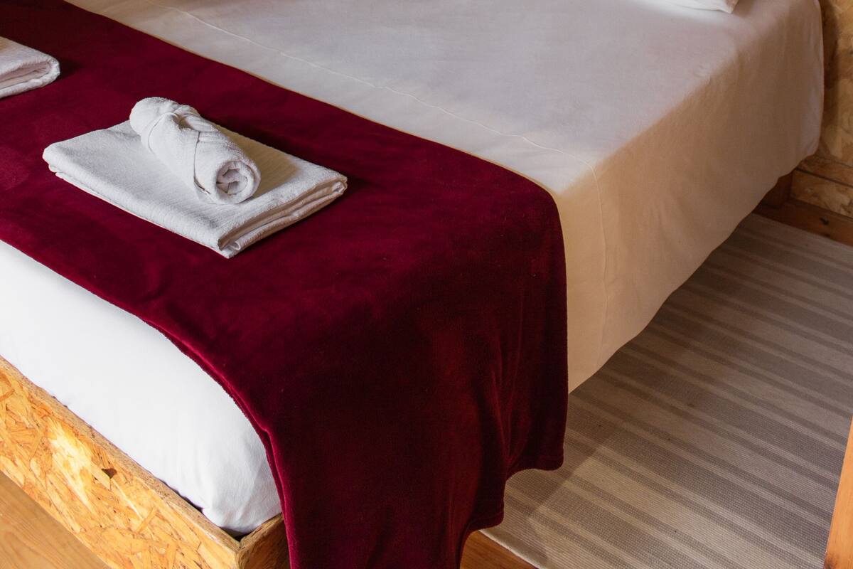 Una habitación con paneles de madera, una cama, ropa de cama blanca, un edredón rojo y una toalla doblada con cuidado.