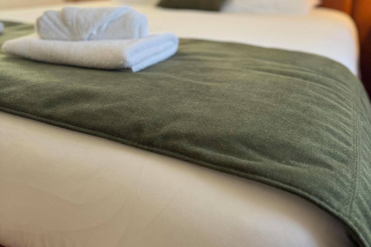 Una cama bien hecha con una manta verde, almohadas blancas y una toalla blanca doblada.
